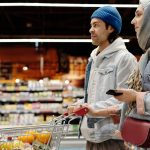 Casal empurrando um carrinho em um supermercado enquanto escolhe frutas e outros alimentos.